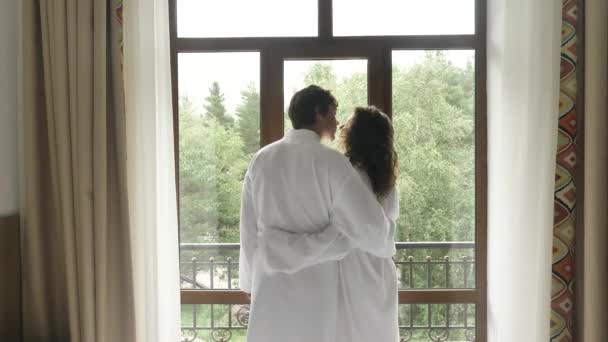 Mladý pár zamilovaný v bílých kabátech se přiblíží k oknu v hotelovém pokoji, políbí, obejme a podívá se na výhled kolem - Záběry, video