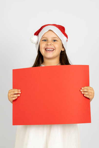 portrait of smiling, funny girl in white dress and Santa Claus hat holding red blank banner with copy space isolated on white background - Foto, Bild