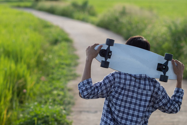 Um jovem segurando um skate de surf em sua mão depois de aperfeiçoar suas habilidades de skate de surf até que ele está cansado e está parando para descansar antes de começar a jogar novamente no caminho para o seu destino pretendido. - Foto, Imagem