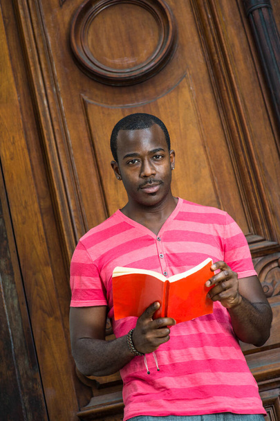 Gekleed in een rood, roze lijnen T-shirt, het dragen van een armband, een jonge zwarte man is het lezen van een rood boek buiten - Foto, afbeelding