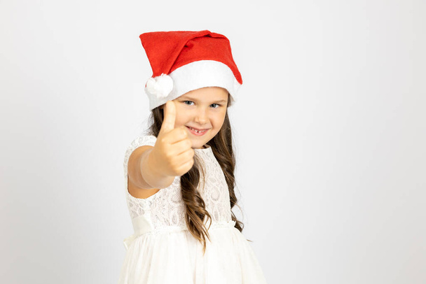 portrait of smiling, enthusiastic child in red Santa Claus hat gives thumbs up, isolated on white background with copy space - Photo, image