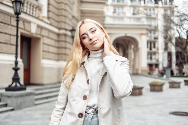 Portret młodej blondynki w jesiennym płaszczu na tle miejskiej architektury - Zdjęcie, obraz