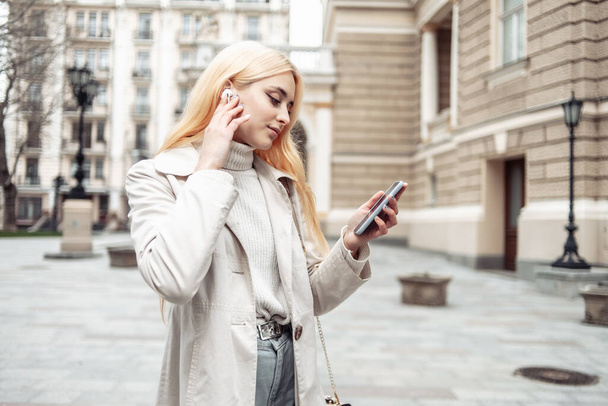 Giovane donna bionda in auricolare utilizzando smartphone in città - Foto, immagini