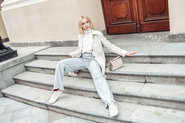 Young fashion blonde woman in a trench coat sits on stairs in the city - Photo, Image