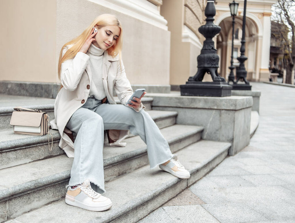 Young fashion woman in a trench coat and jeans sits on the stairs and uses a smartphone - Photo, Image