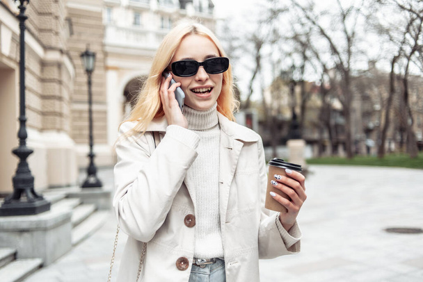Güneş gözlüklü genç sarışın kadın telefonda konuşuyor ve şehirde elinde bir fincan kahve tutuyor. - Fotoğraf, Görsel