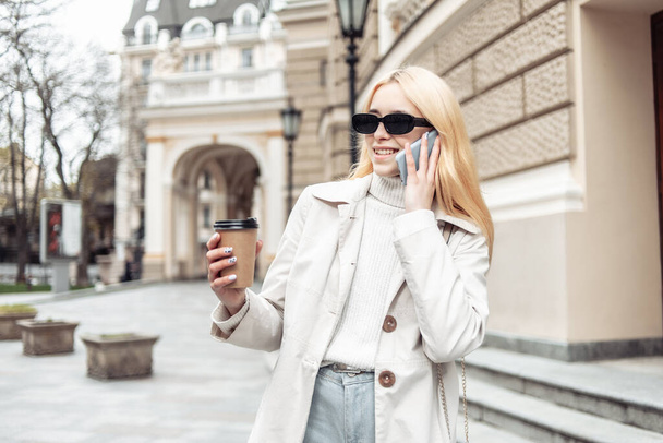 Młoda blondynka w okularach przeciwsłonecznych rozmawia przez telefon i trzyma filiżankę kawy w mieście - Zdjęcie, obraz