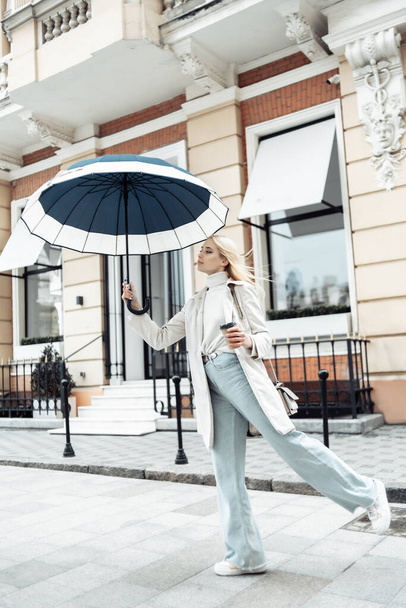 Молодая блондинка в пальто с зонтиком и чашкой кофе в городе - Фото, изображение