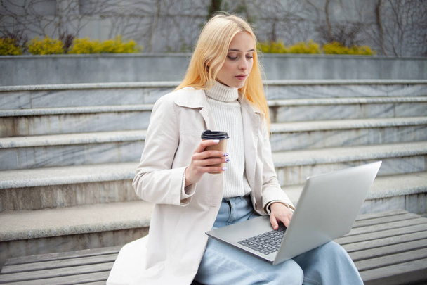 Young woman student uses a laptop and drink coffee while sitting on bench - Photo, Image