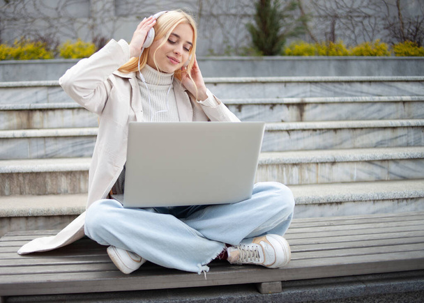 Молодая веселая женщина слушает музыку в наушниках, сидя на скамейке с ноутбуком - Фото, изображение