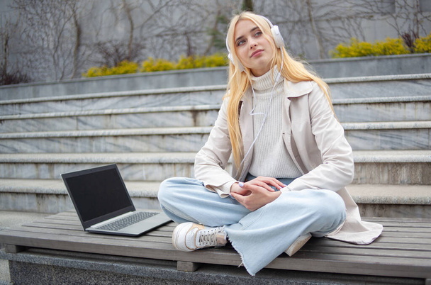 Young cheerful woman student listening to music in headphones while sitting on a bench with laptop - Photo, Image