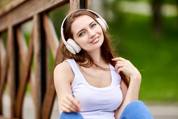 Smiling young woman in wireless headphones sits outdoors. - Fotó, kép