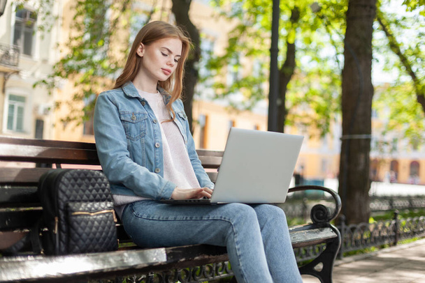 Young pretty woman student or freelancer using laptop while sitting on a bench in the city - Zdjęcie, obraz