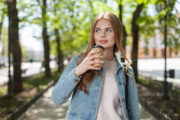 Fiatal fehér nő farmerdzsekiben kávét iszik útközben a városban. - Fotó, kép