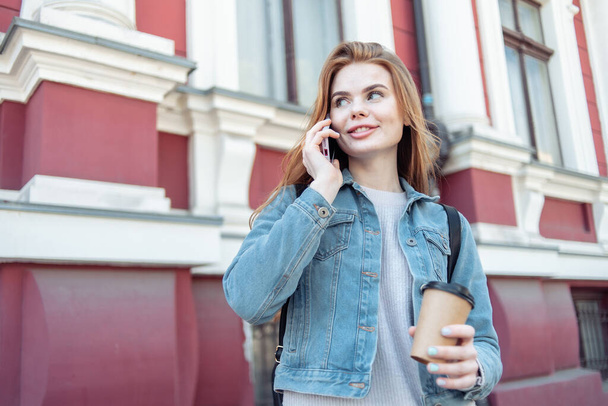 Νεαρή χαμογελαστή γυναίκα που μιλάει στο τηλέφωνο στην πόλη και κρατάει ένα φλιτζάνι καφέ  - Φωτογραφία, εικόνα