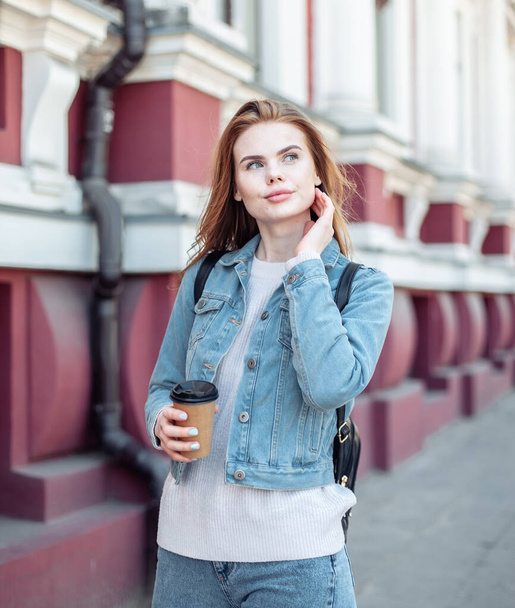 Νεαρή όμορφη ελκυστική γυναίκα με τζιν μπουφάν που κρατάει μια κούπα καφέ στην πόλη. - Φωτογραφία, εικόνα