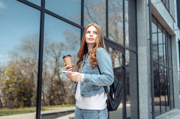 Young beautiful woman in denim jacket holding a cup of coffee near the windows of the business center - Photo, image