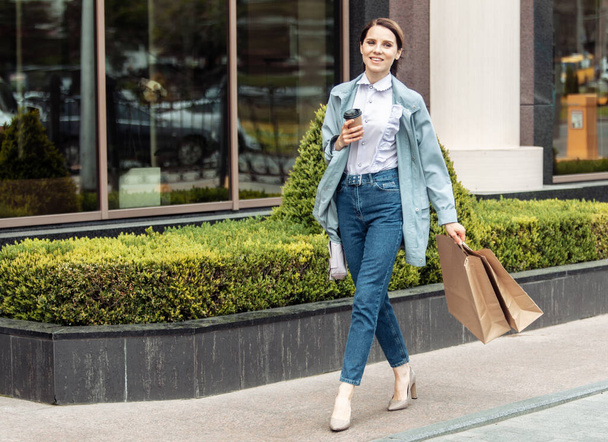 Pretty, cheerful lady is walking along city street with shopping bags and drinking coffee on the go. Lifestyle shopping concept - Photo, Image