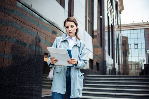 Aufmerksam blickt die moderne Geschäftsfrau in Ordner mit Dokumenten vor dem Hintergrund eines Geschäftshauses mit Treppenhaus. Lebensstil - Foto, Bild