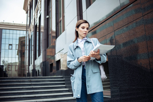 Modern iş kadını, iş binasının arka planında merdiveni olan belgeleri dikkatle inceliyor. Yaşam biçimi - Fotoğraf, Görsel