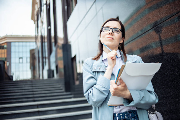 Pensive young business woman in glasses with a folder of documents in the city. Lifestyle - 写真・画像