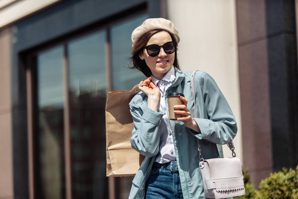 Femme commerçante élégante en trench-coat tendance, béret, lunettes de soleil en ville avec des sacs à provisions et une tasse de café. Concept de shopping. Mode de vie - Photo, image