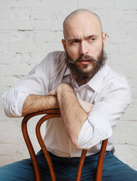 portrait, white young bald man with a beard and mustache in a white shirt sitting on a chair in the room,  - Foto, imagen