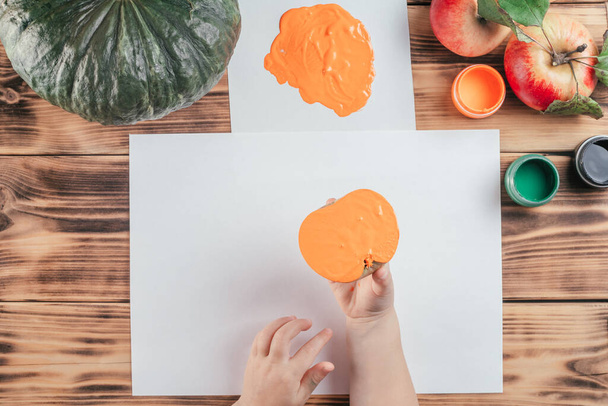 Step-by-step Halloween childrens tutorial pumpkin apple prints. Step 7: Child hand holds half of apple painted in orange gouache to make print. Top view - Photo, Image