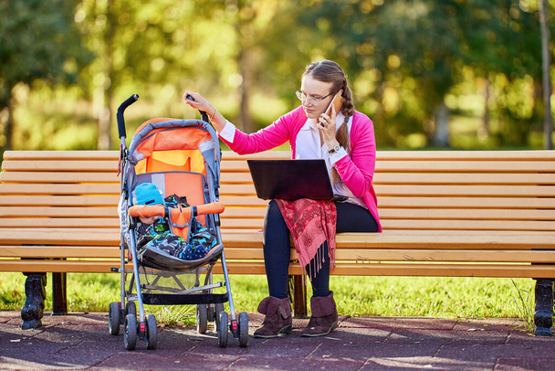 Woman with infant in pram is talking by phone outdoors. - Фото, изображение