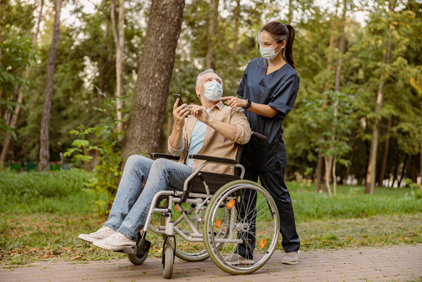 Jovem enfermeira no escudo facial e máscara protetora assistindo o homem deficiente sênior em cadeira de rodas usando seu smartphone durante uma caminhada no parque - Foto, Imagem