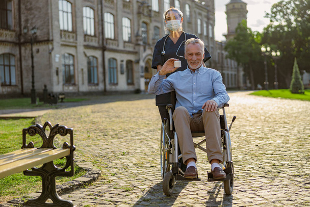 In voller Länge Aufnahme eines glücklichen älteren Mannes, der sich bei einem Spaziergang im Freien in der Nähe einer Reha-Klinik von einem Patienten mit einem Cremeglas erholt und in die Kamera lächelt - Foto, Bild