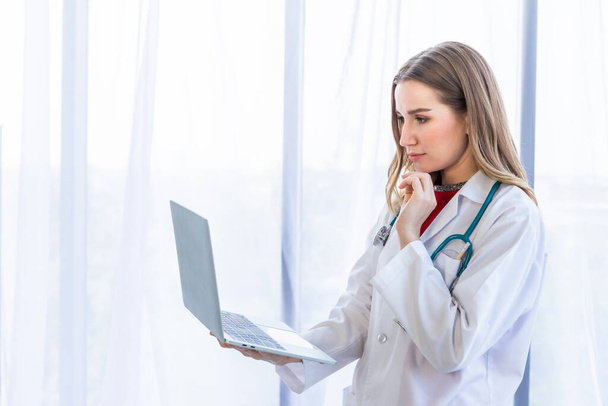 femme médecin avec stéthoscope travaillant avec ordinateur portable et écriture sur la paperasserie sur table en bois dans le fond de l'hôpital - Photo, image