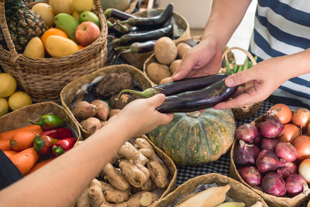 Ein Mann reicht einem Verkäufer zwei Auberginen seiner Wahl. Verschiedenes Gemüse und Obst im Hintergrund. Ein Kauf an einem kleinen Marktstand. - Foto, Bild