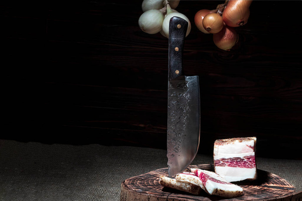 dark background. round wooden tub in retro style. a piece of bacon is cut into pieces. a sharp knife made of forged steel. black bread. - Photo, Image