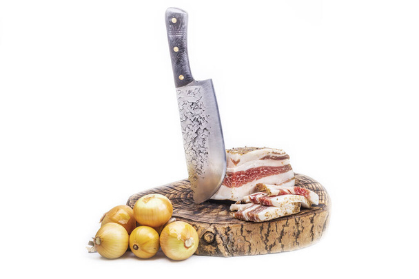 wooden stand. a sharp knife in retro style. a piece of delicious bacon and a slice is cut next to it. the golden yellow heads of the bulbs lie next to each other. on a white background. - Photo, Image