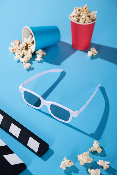 Fehér 3D-s filmszemüveg, popcorn papírpohárban, tapsoló tábla kék háttérrel. Mozi szórakoztató koncepció. függőleges fénykép. - Fotó, kép