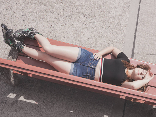 Happy joyful young woman wearing roller skates lying on bench with raised legs. Bizarre high angle - 写真・画像