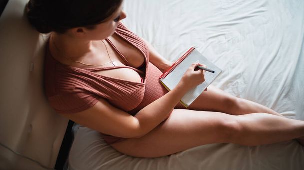 Pregnant girl sits on a bed and irons her stomach with a notebook on her lap. List to the maternity hospital. Waiting for the baby. Place for text. Copy space - Fotografie, Obrázek