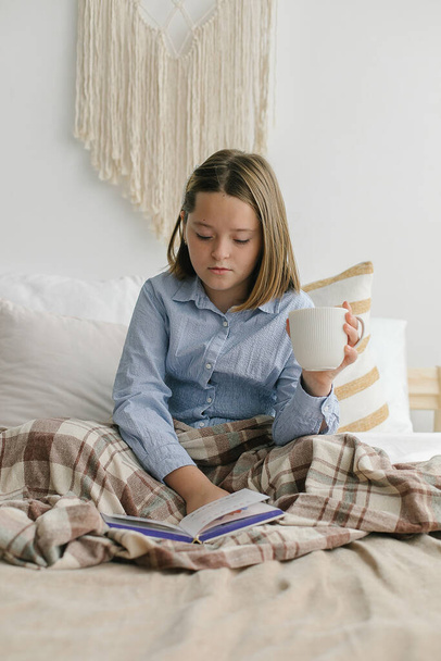 Девушка с чашкой чая или шоколада, читает книгу, отдыхает, сидя на кровати. Уютный сезон дома, гигге, комфорт, настроение концепции - Фото, изображение