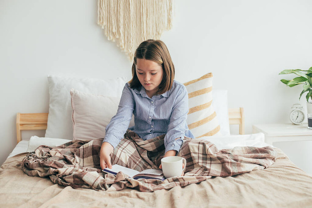 Chica con taza de té o chocolate, leer un libro, descansar mientras está sentado en la cama. Acogedora temporada en casa, hygge, comodidad, concepto de estado de ánimo - Foto, Imagen