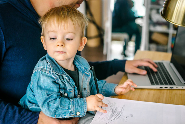 Babası küçük oğlunu kucağına alıp laptopla evde çalışıyor. Uzaktan çalışma kavramı - Fotoğraf, Görsel