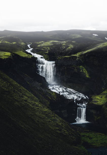 A mesmerizing vertical shot of a river with waterfalls flowing over the green mountains - Фото, изображение