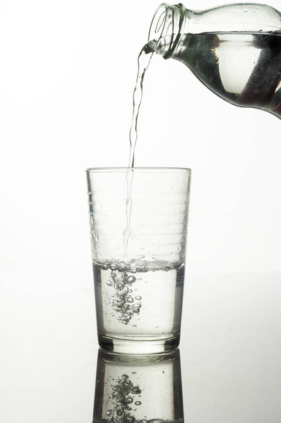 Вертикальный снимок процесса разлива воды из бутылки в стакан на белом фоне - Фото, изображение