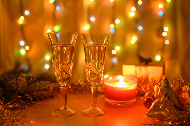 Ποτήρια αφρώδους κρασιού περιτριγυρισμένα από την Πρωτοχρονιά με ένα αναμμένο κερί και μια φωτεινή γιρλάντα. Ρηχό βάθος πεδίου. - Φωτογραφία, εικόνα