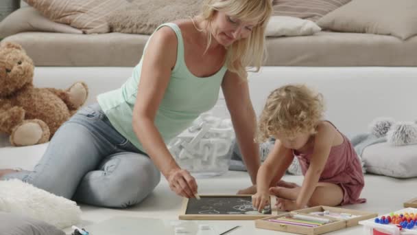 glimlachende moeder lacht naar krullend blond dochter kind, kleuterschool leeractiviteit thuis, tekent en schrijft spelen met krijt op schoolbord in zonnige woonkamer, concept van gezond groeien - Video