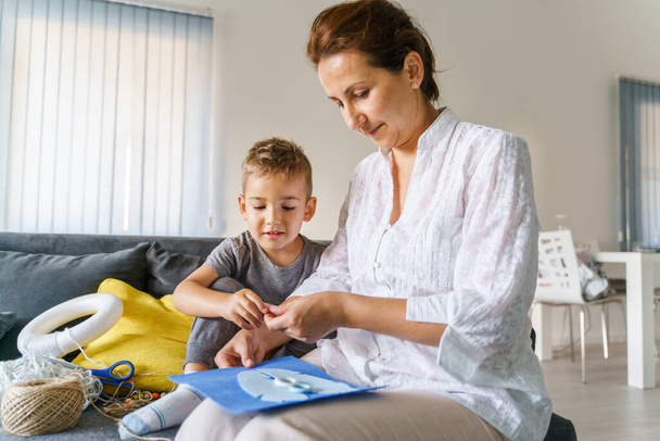Small caucasian boy having fun with his mother making homemade decoration - Little child son with mature woman on the sofa at home playing - hobby creativity development real people childhood concept - Photo, Image