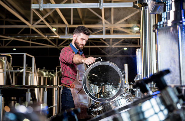 Jeune brasseur masculin en tablier de cuir supervisant le processus de fermentation de la bière à l'usine de brasserie moderne - Photo, image