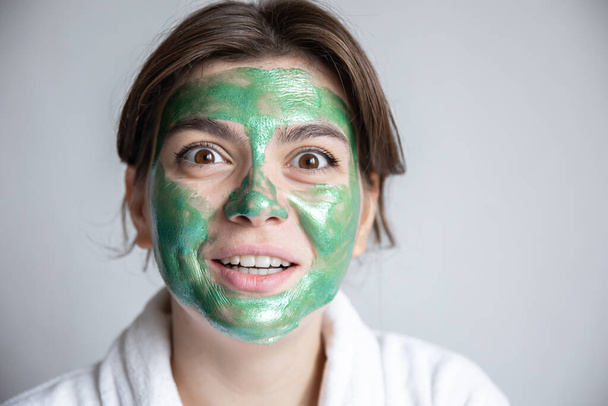 Ελκυστική νεαρή γυναίκα με πράσινη καλλυντική μάσκα στο πρόσωπό της και με λευκή ρόμπα σε γκρι φόντο, η έννοια των θεραπειών spa στο σπίτι, αντιγραφή χώρου. - Φωτογραφία, εικόνα