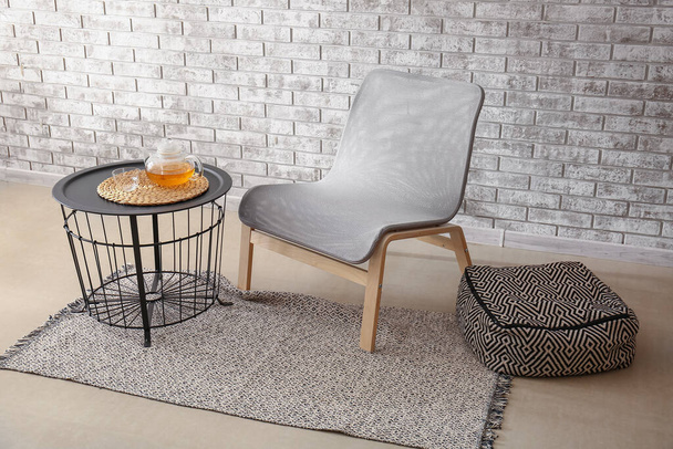 Gezellige fauteuil, poef en tafel met theepot bij bakstenen muur - Foto, afbeelding