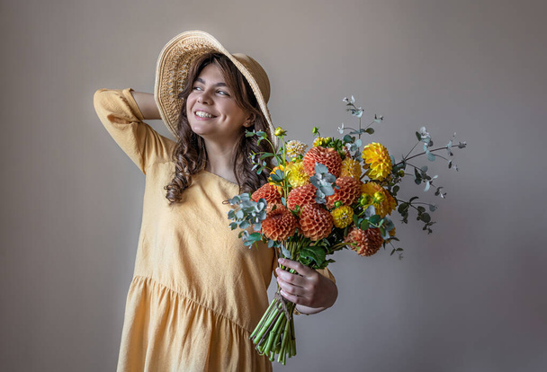 Fröhliche junge Frau in gelbem Kleid und Hut mit einem Strauß heller Chrysanthemen auf grauem Hintergrund. - Foto, Bild